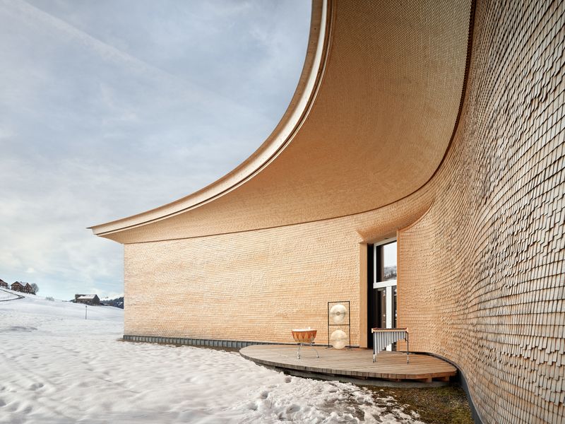 Klanghaus Toggenburg am Schwendisee – Parbolisch geformter Aussenraum mit vorgelagerter Klang- bzw. Hörbühne und «visueller Akustik» in der Schindelverkleidung. Im Laufe der Zeit wird sich unter den ausladend gekrümmten Vordächern durch die Verwitterung eine zweite Wellenform über die wellenförmige Ornamentik bei den Aussenbühnen legen. Der exponierte Bereich des Schindelschirms dunkelt schneller nach als der geschützte. – Roland Bernath